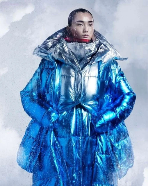 Asian adult model wearing a metallic blue oversized puffer jacket with an exaggerated collar against a blurred cloudy background.