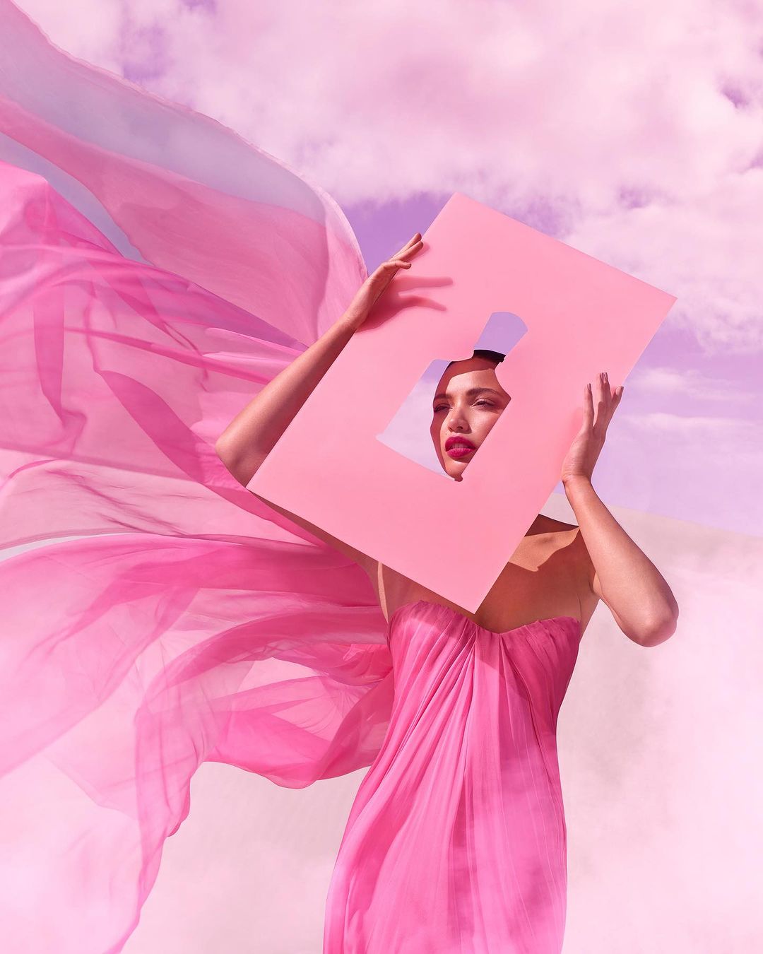 Woman in a pink dress holding a pink frame with a perfume bottle cutout, set against a pink cloudy sky and desert-like terrain.