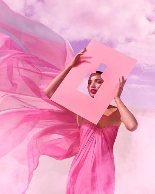 Woman in a pink dress holding a pink frame with a perfume bottle cutout, set against a pink cloudy sky and desert-like terrain.
