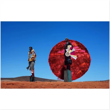 Two women stand tall on wooden platforms in a sunny desert landscape under a clear blue sky, showcasing fashion and style.