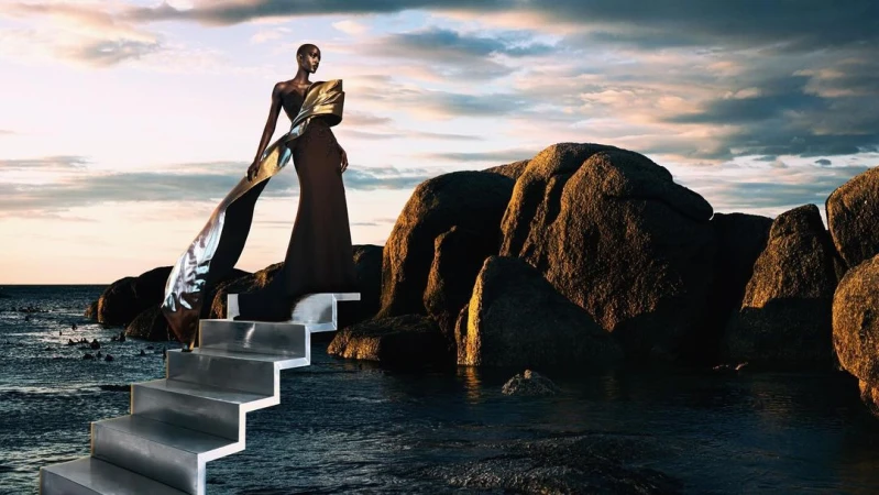 A bald Black woman wearing a structured dark gown with a metallic gold sash standing on metallic stairs by rocky coast and ocean at sunset.