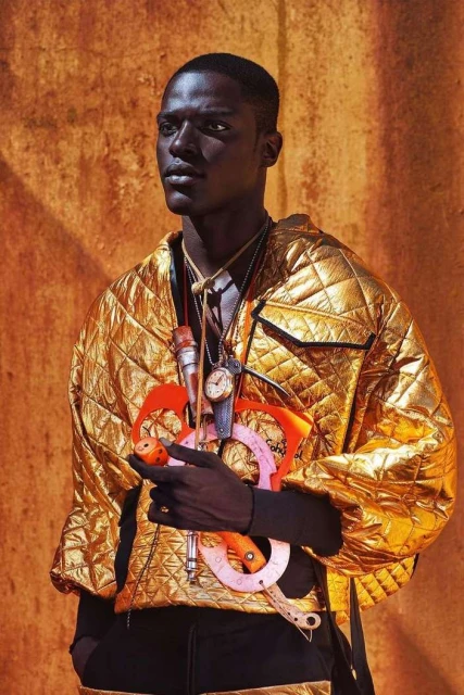 Stylish Black man poses in a gold quilted jacket, accessories, against a textured orange wall in a fashion editorial shot.