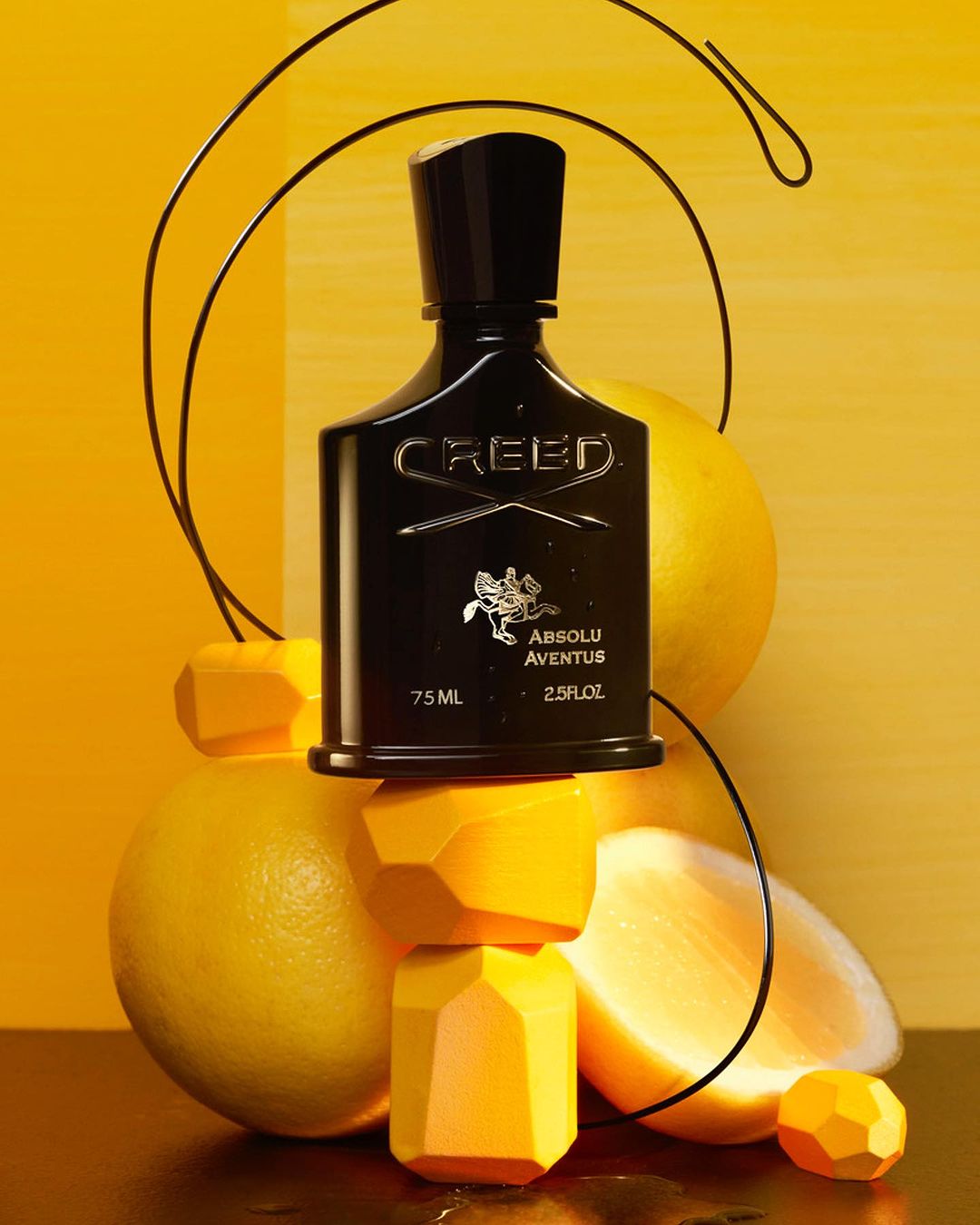 Creed Absolu Aventus perfume bottle sits atop yellow geometric blocks and lemons against a split yellow backdrop with black wire.