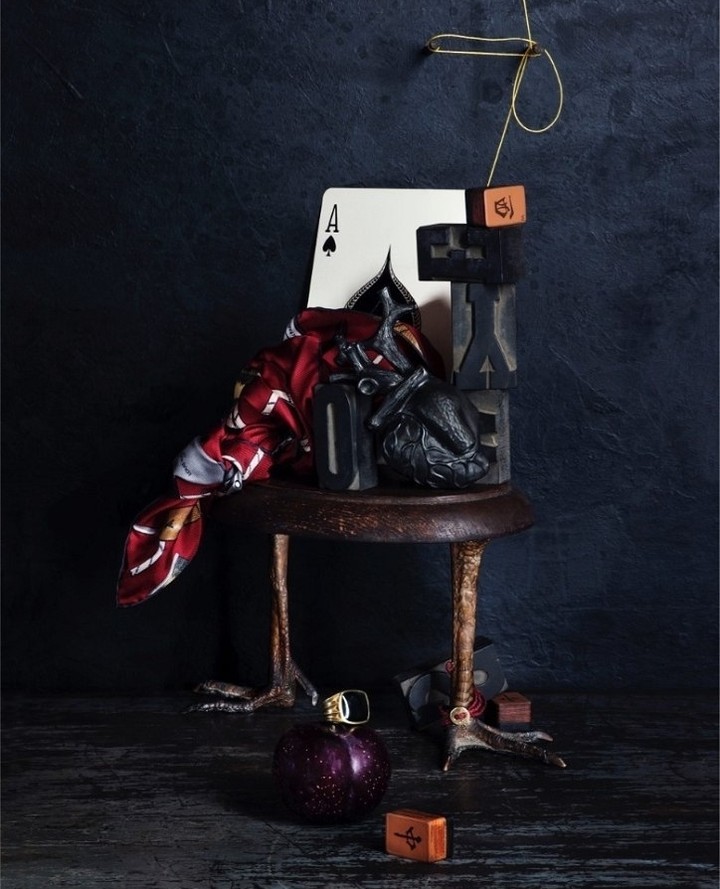 Still life with an Ace of Spades card, sculpted crow, decorative objects on a small table with bird-like legs, and dark background.