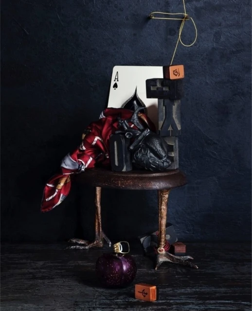Still life with an Ace of Spades card, sculpted crow, decorative objects on a small table with bird-like legs, and dark background.