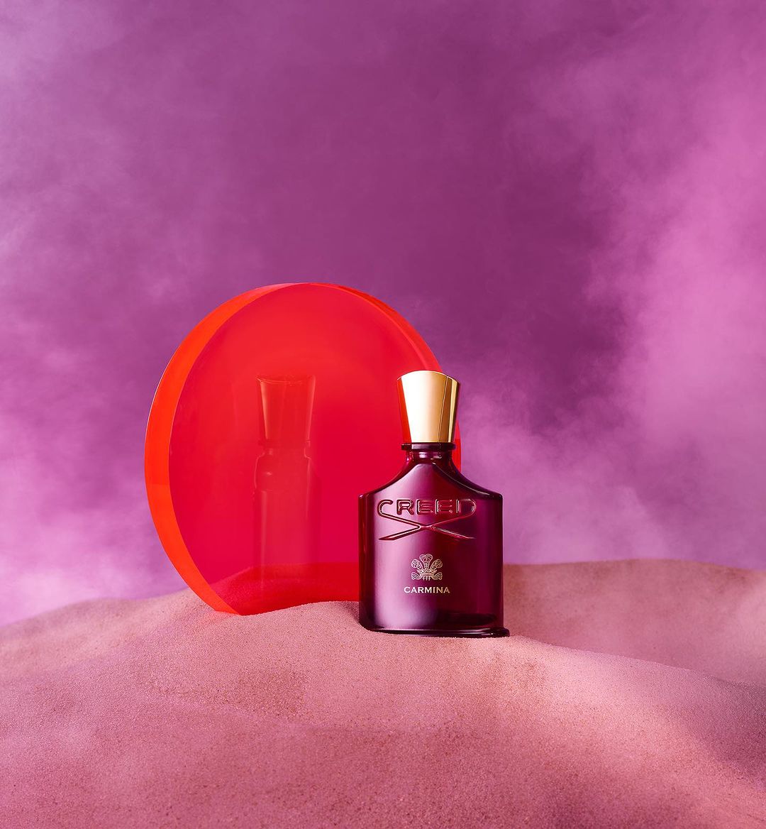 Creed Carmina perfume bottle with a gold top on a pink surface with a red transparent circle and purple background.