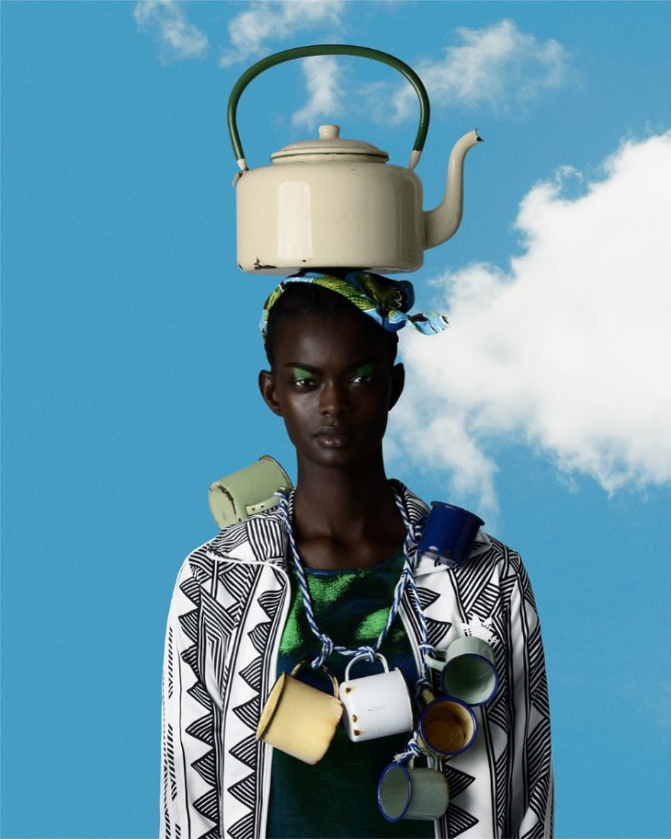 Portrait of an African woman with a teapot on her head and mugs around her neck, posing in front of a cloudy blue sky.
