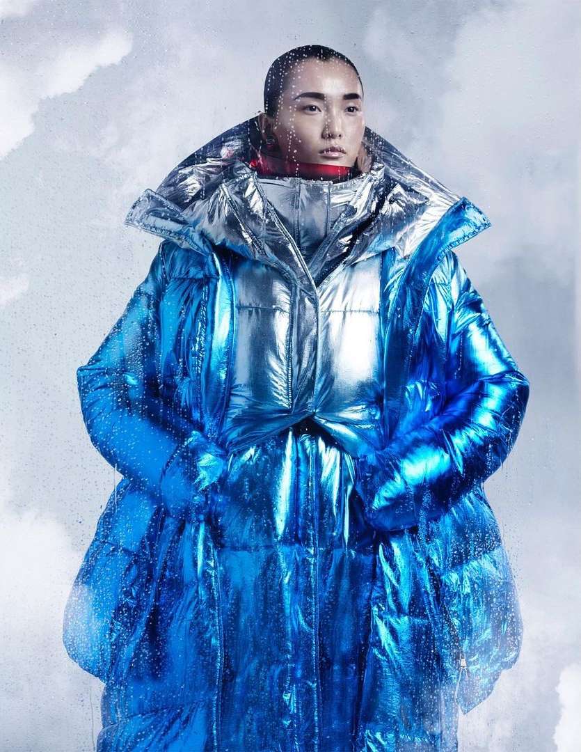 Woman in iridescent blue puffer coat poses against a cloudy background in a studio fashion shot with creative lighting.