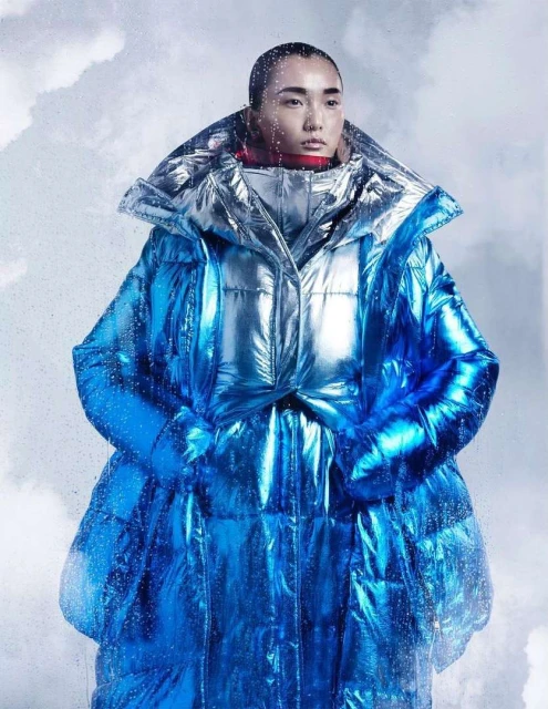 Woman in iridescent blue puffer coat poses against a cloudy background in a studio fashion shot with creative lighting.