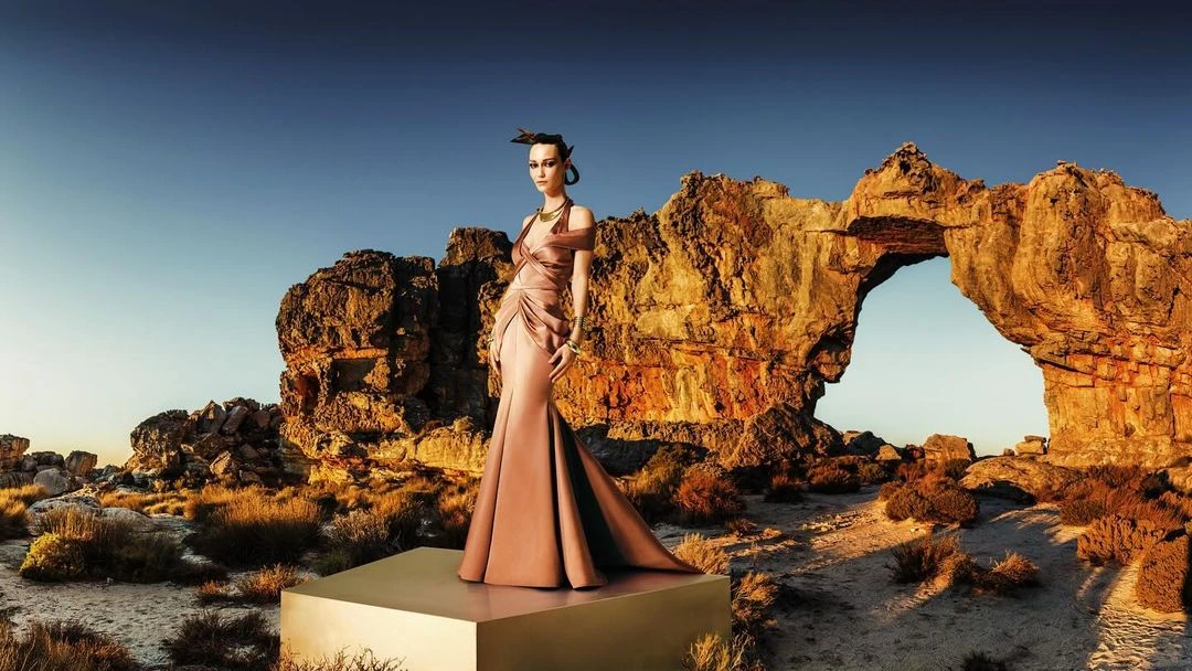Fashion model in a long flowing dress strikes a pose atop a metallic platform in a rocky, natural landscape with a blue sky backdrop.