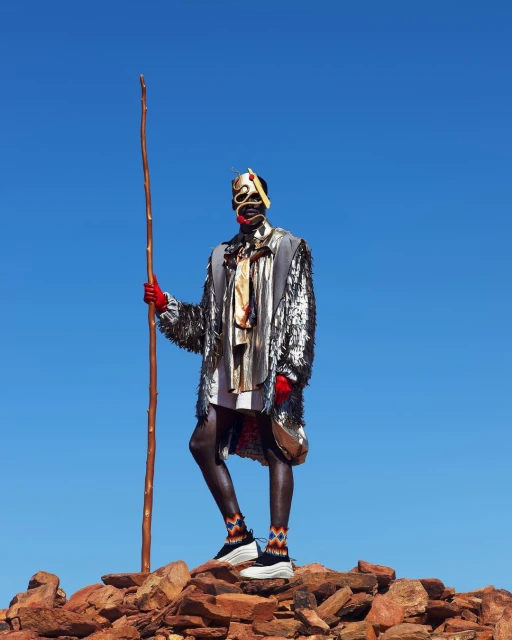 Person in patterned mask, sparkling silver coat, and patterned socks standing on a rocky hilltop against a clear blue sky holding a wooden stick.