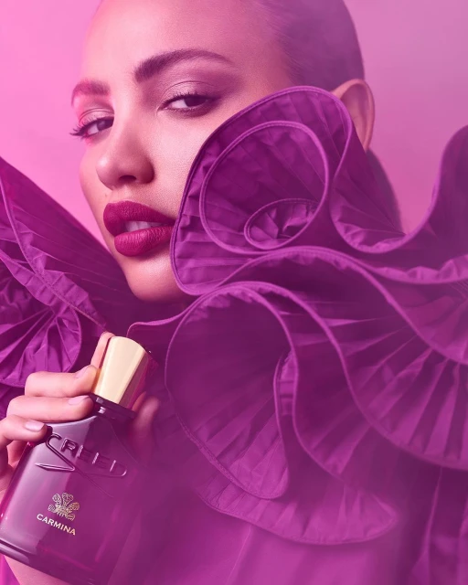 Close-up of a brunette adult female holding a Carmina perfume bottle, wearing a magenta ruffled top against a pink gradient backdrop.