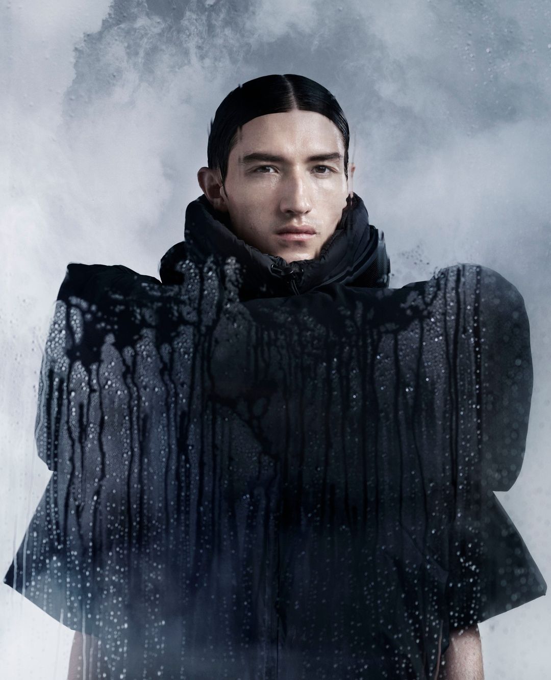 Portrait of a male fashion model in a black hooded jacket with water droplet details against a cloudy grey background, studio shot.