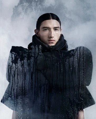 Portrait of a male fashion model in a black hooded jacket with water droplet details against a cloudy grey background, studio shot.