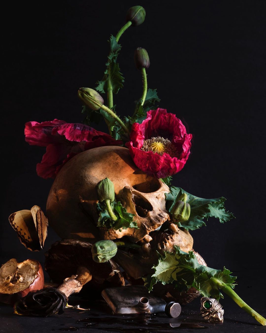 Skull still life with red poppy flowers, green leaves, vintage bottle, jewelry, and apple on a dark background. Gothic-inspired art.