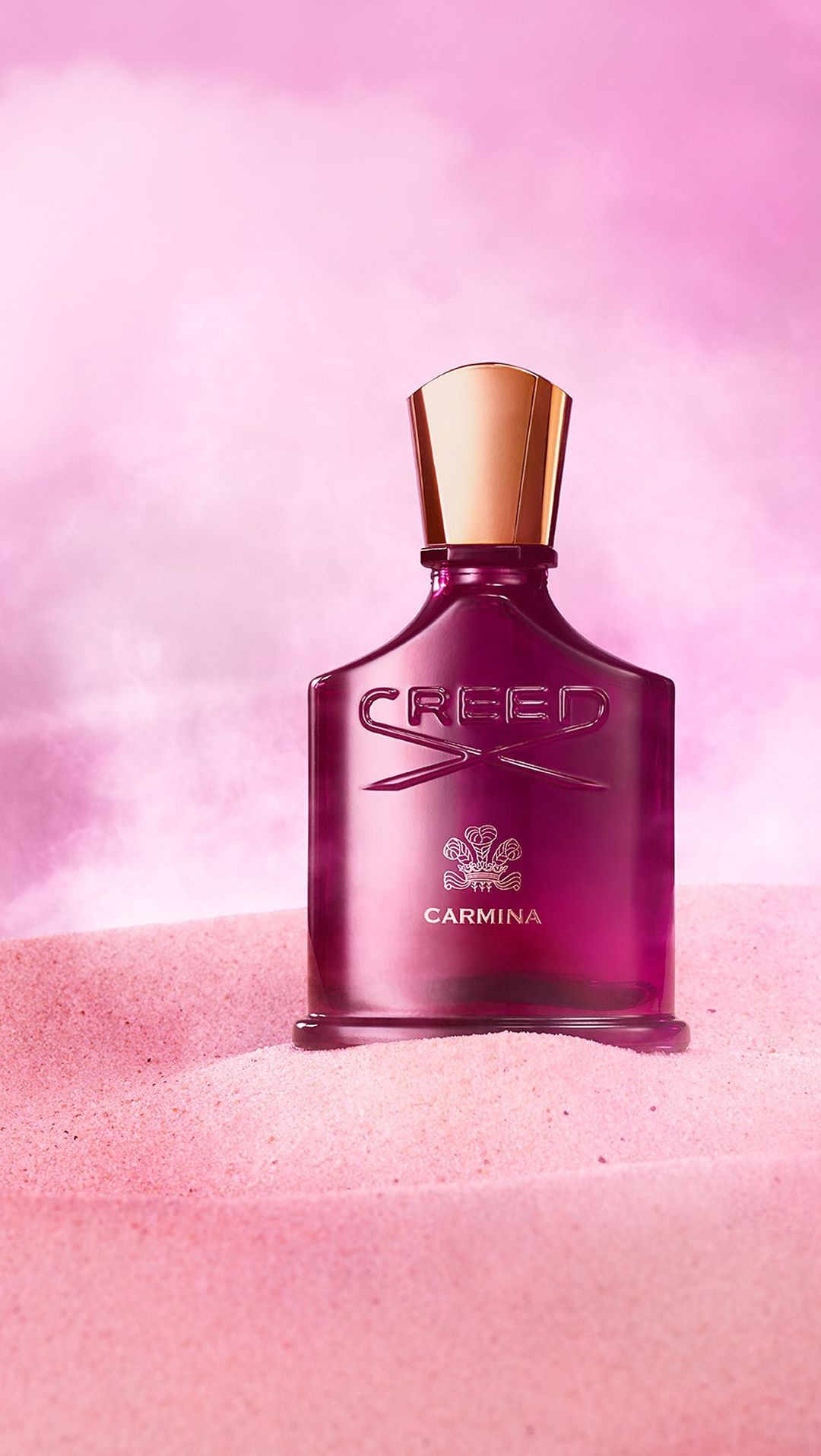 Close-up studio shot of Creed Carmina perfume bottle with gold cap on pink sand, against a pink and white cloud-like backdrop.