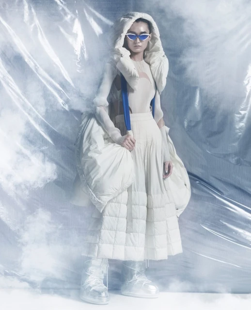 Fashion shot of a woman in a white hooded jacket, cream dress, and shiny boots, standing in front of a wrinkled plastic backdrop.