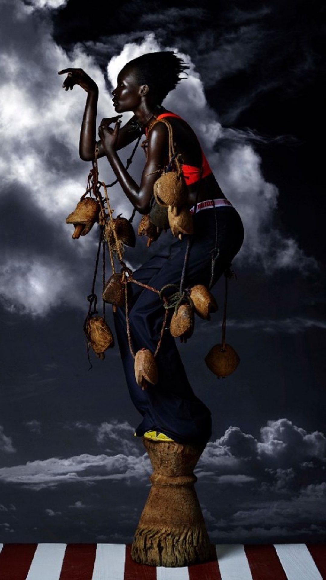 Fashion shot of a black female model on a pedestal wearing a red top and dark pants, holding coconuts on a string, against a cloudy sky.