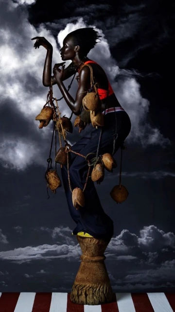 Fashion shot of a black female model on a pedestal wearing a red top and dark pants, holding coconuts on a string, against a cloudy sky.