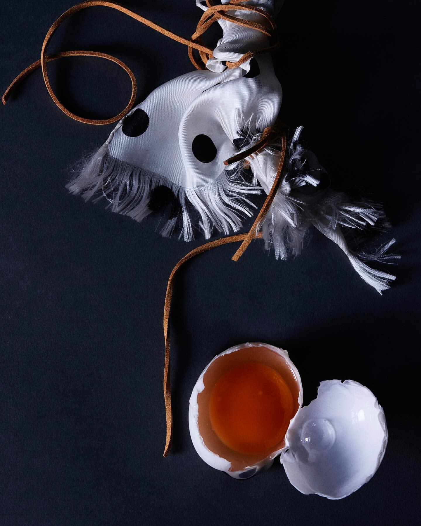 Still life photograph featuring a cracked egg with yolk, a white scarf with black polka dots, and a brown leather string on a dark surface.