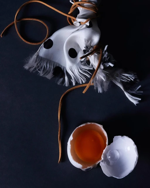 Still life photograph featuring a cracked egg with yolk, a white scarf with black polka dots, and a brown leather string on a dark surface.