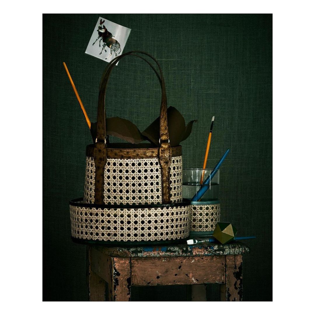 Still life composition featuring a woven basket-style handbag, glass with water and paintbrush, atop a distressed wooden stool against a dark green background.