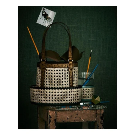 Still life composition featuring a woven basket-style handbag, glass with water and paintbrush, atop a distressed wooden stool against a dark green background.