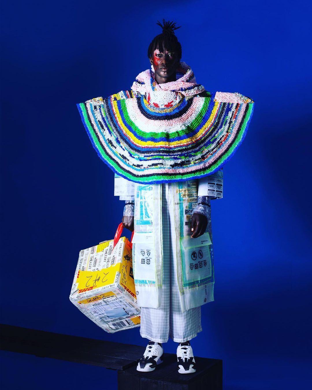 Person in avant-garde fashion stands on platform wearing coat and head covering made of recycled materials with blue background.