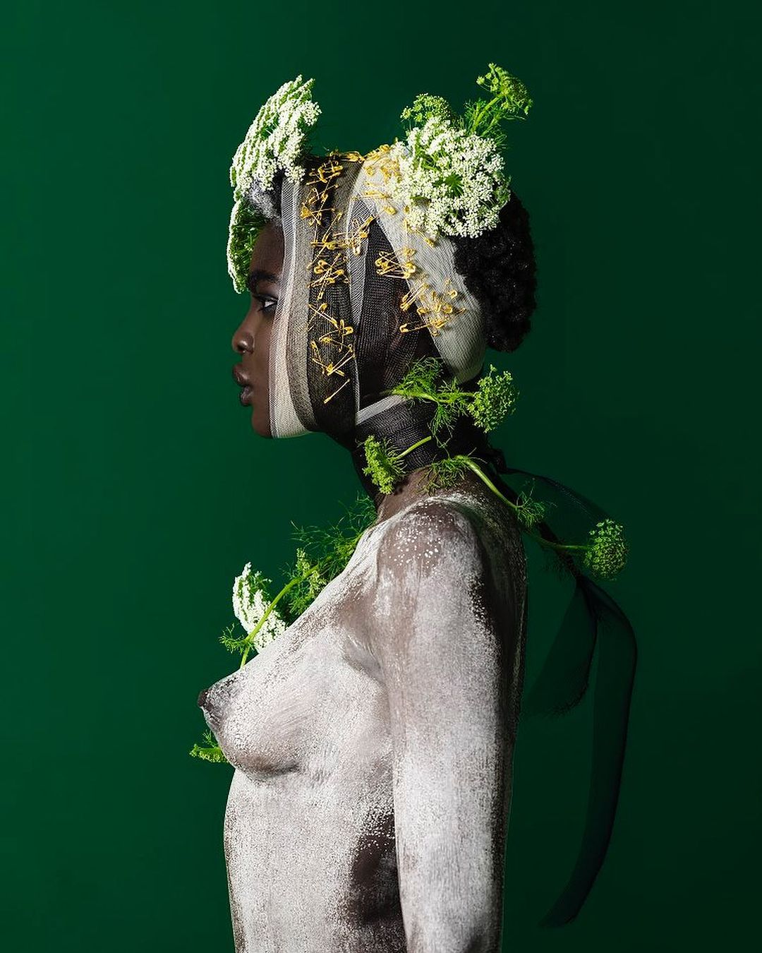 A woman with dark skin is painted white and adorned with white flowers and green foliage, captured in a studio shot against a green backdrop.
