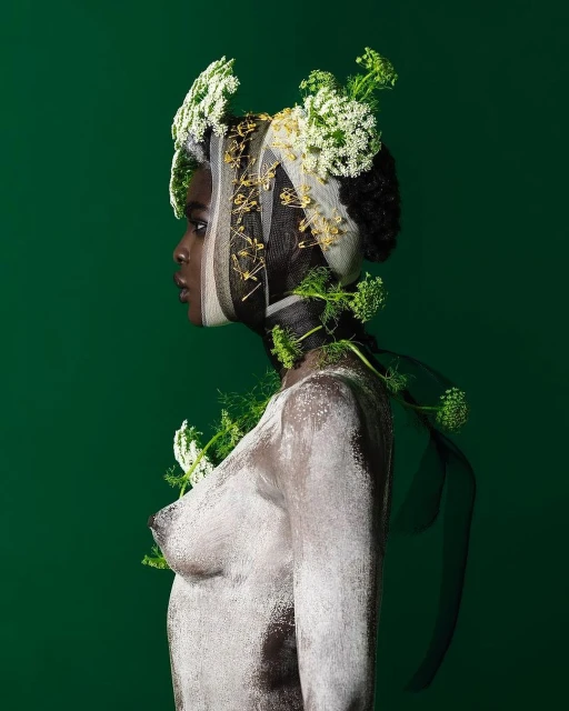 A woman with dark skin is painted white and adorned with white flowers and green foliage, captured in a studio shot against a green backdrop.