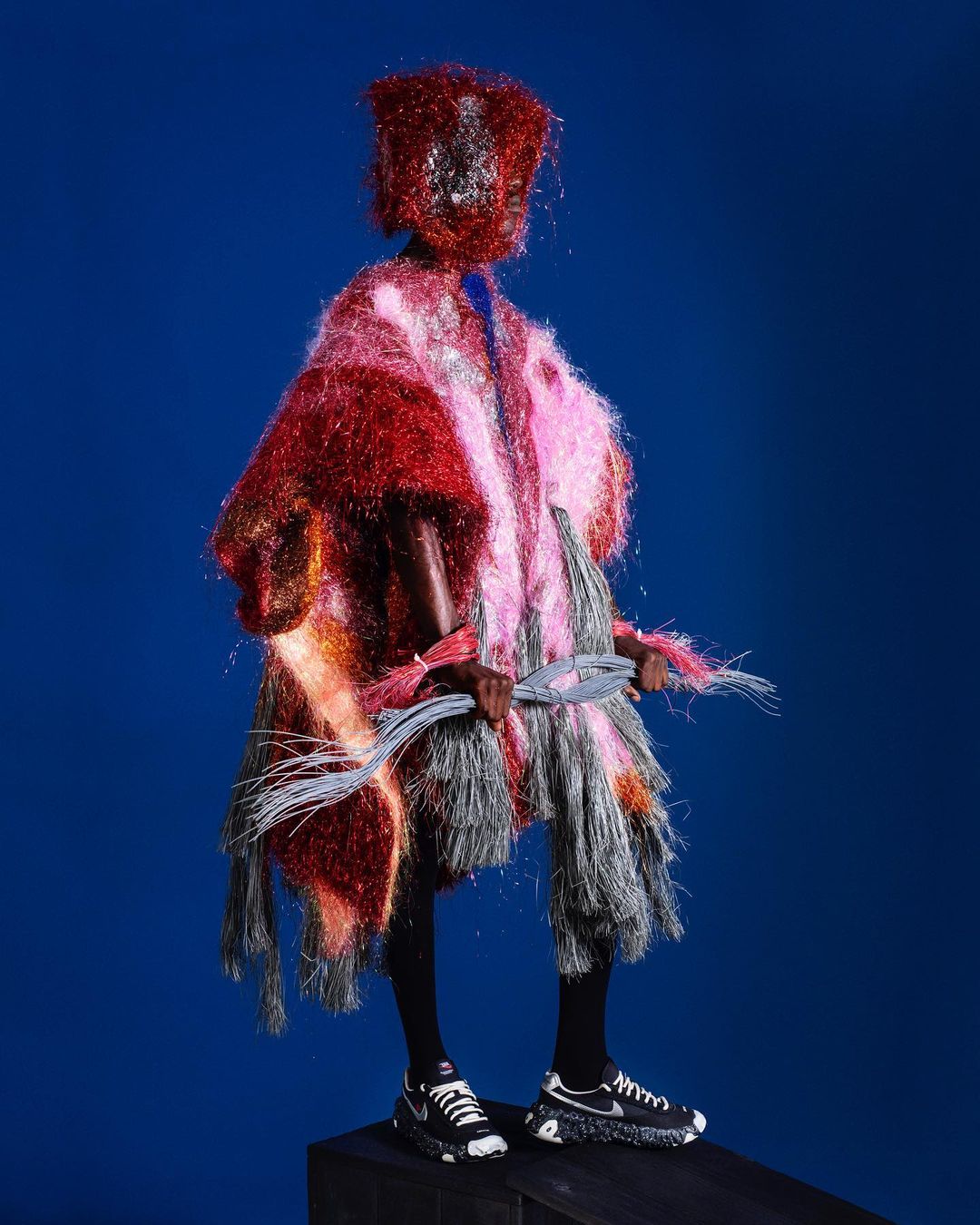 Person in a vibrant textured poncho, head covering, and black platform sneakers stands on a black block against a blue backdrop.