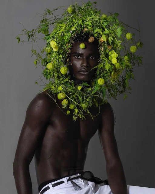 Striking image of a shirtless African male model adorned with vibrant green plants against a gray backdrop, showcasing unique fashion.