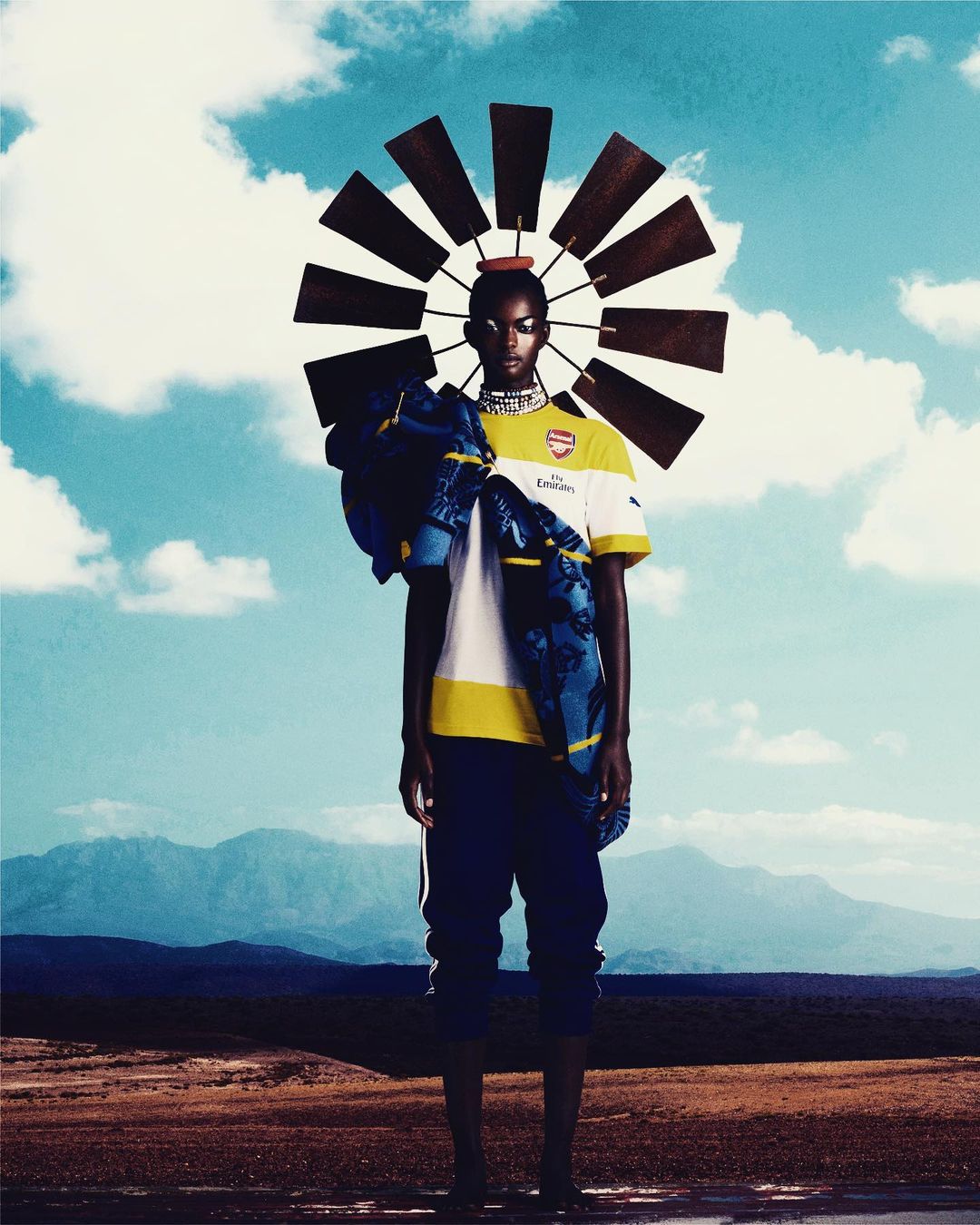 Woman in soccer jersey stands in a desert landscape beneath a vintage windmill headpiece against a partly cloudy blue sky.