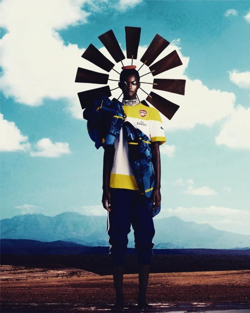Woman in soccer jersey stands in a desert landscape beneath a vintage windmill headpiece against a partly cloudy blue sky.