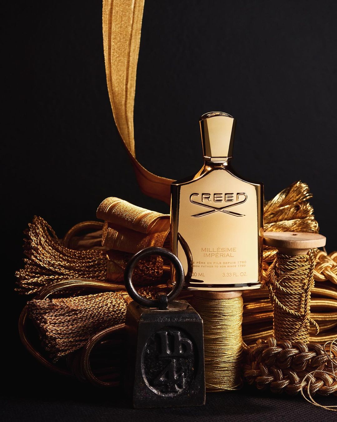 Gold Creed perfume bottle stands out against a black background, surrounded by gold cords, threads, and tassels creating a luxurious feel.
