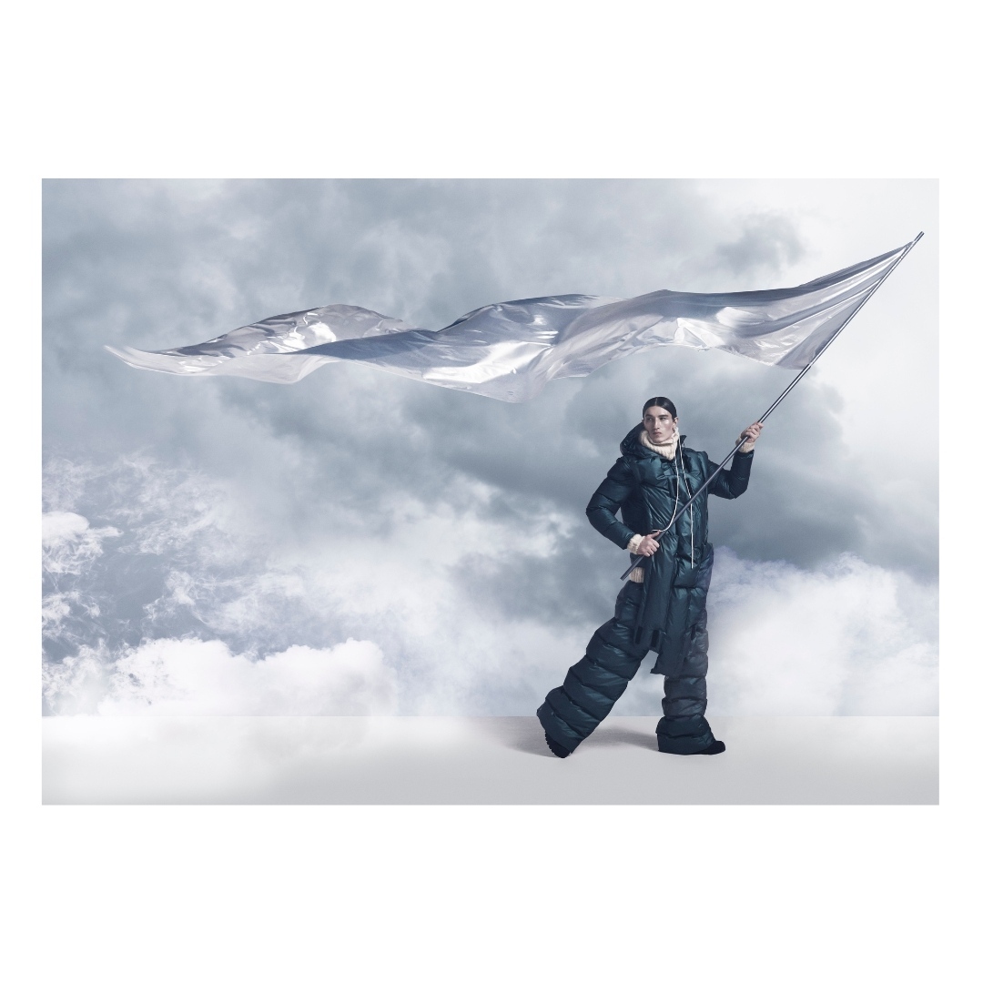 A man in a padded suit stands on a white surface holding a large, silver, flowing flag against a background of wispy clouds.