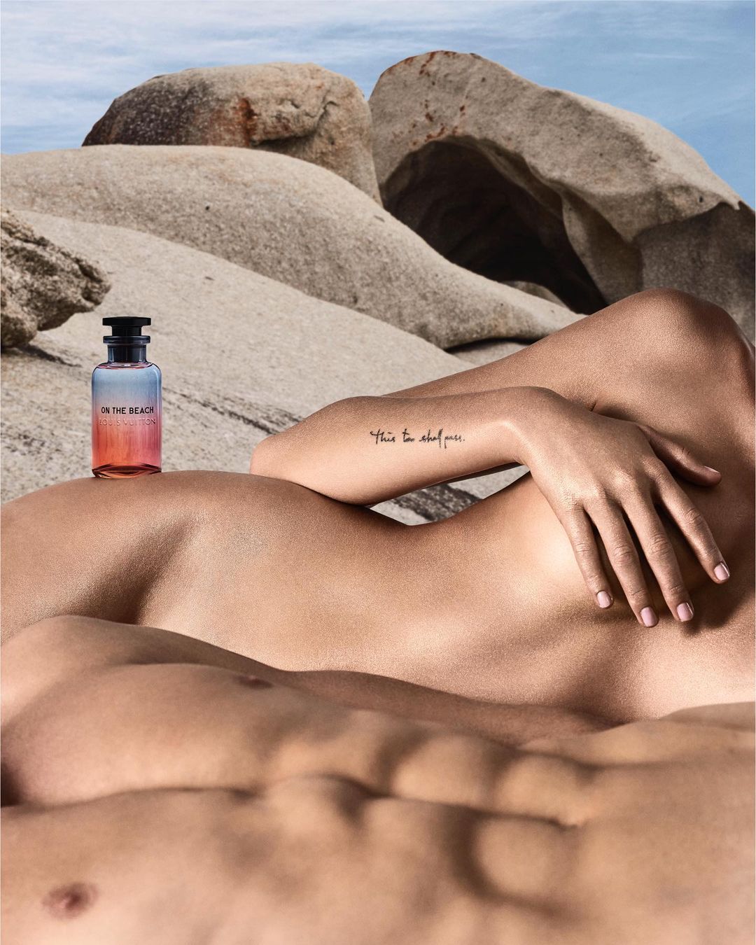Product shot of a perfume bottle resting on a couple on a beach with large rock formations in the background on a bright day.