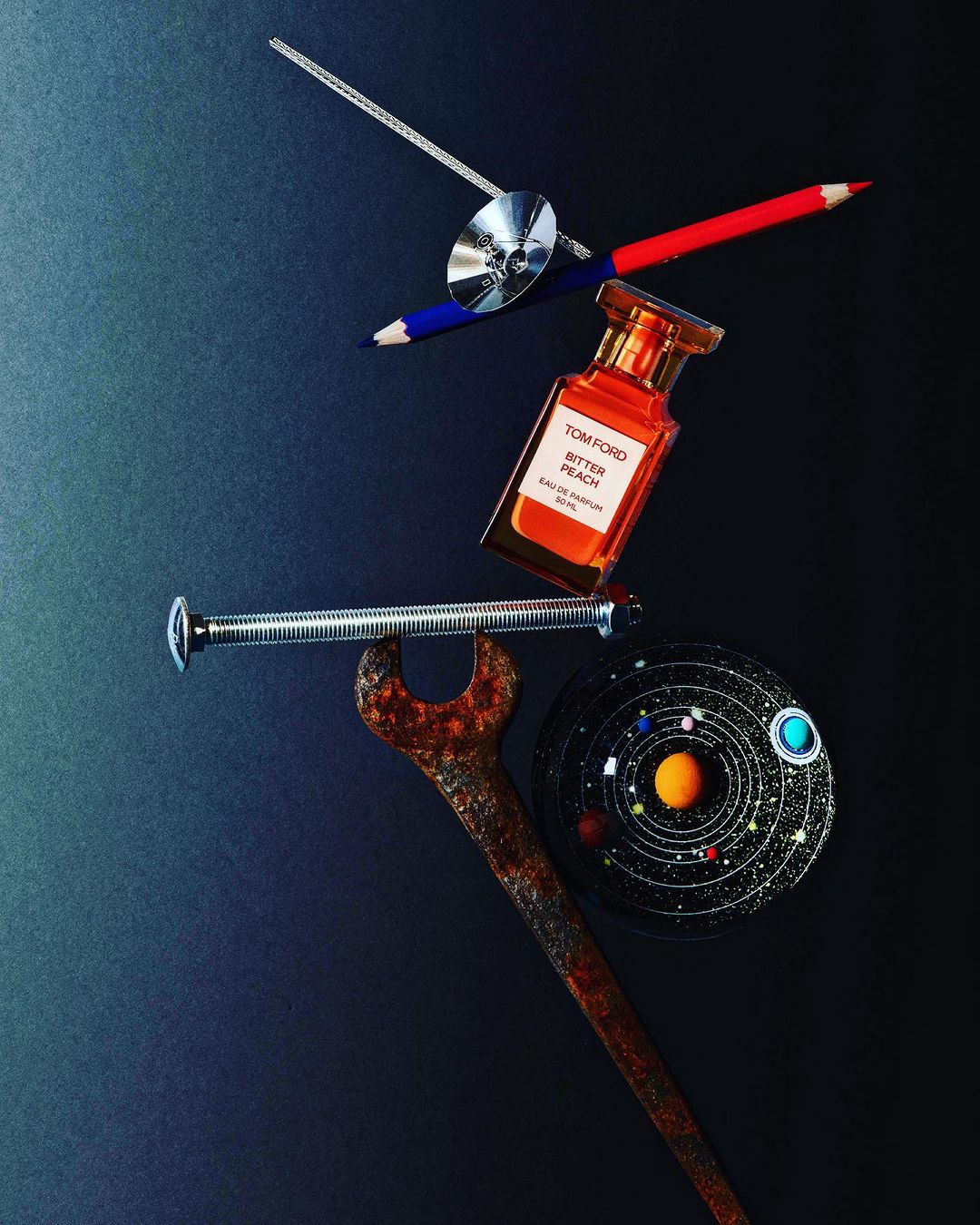 Still life flat lay featuring a perfume bottle, colored pencils, a wrench, a metal screw, and a cosmic art piece on a dark background.