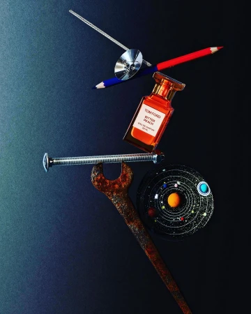 Still life flat lay featuring a perfume bottle, colored pencils, a wrench, a metal screw, and a cosmic art piece on a dark background.