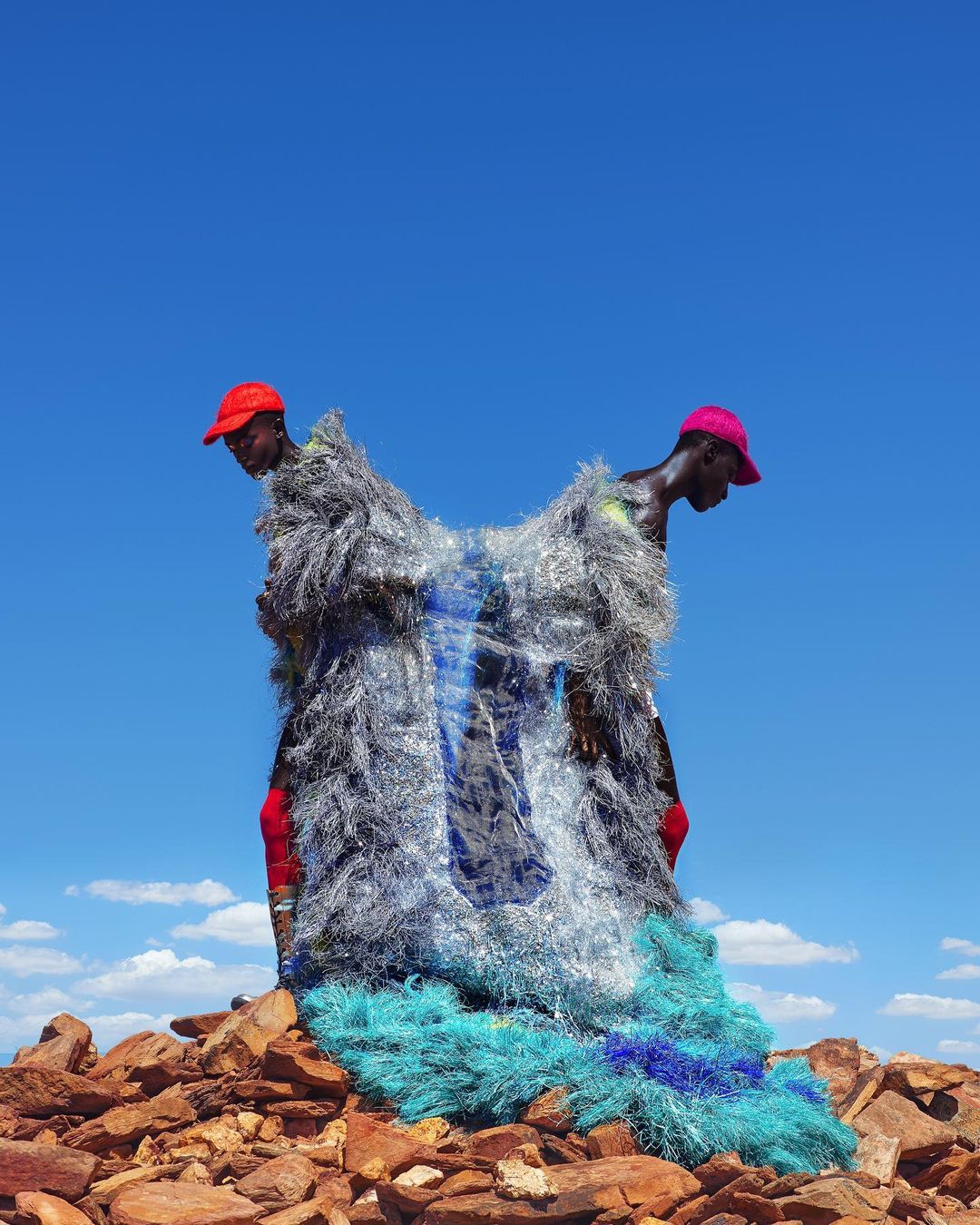Two African men stand on a pile of rocks against a bright blue sky, adorned in a shaggy, shimmering costume with pops of blue.