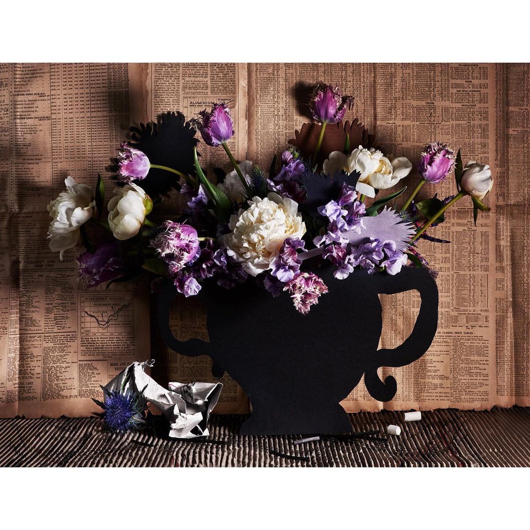Floral arrangement with purple and white flowers in a black vase against a backdrop of vintage newspaper and striped surface.