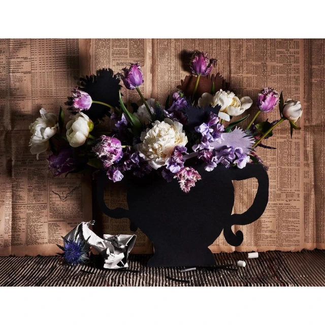 Floral arrangement with purple and white flowers in a black vase against a backdrop of vintage newspaper and striped surface.