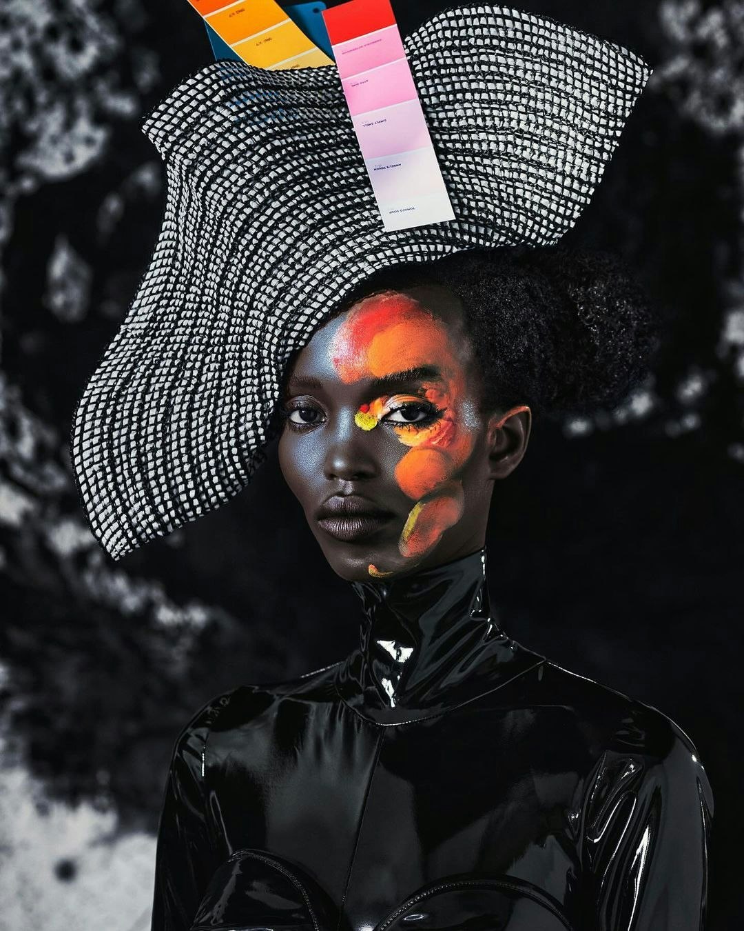 Artistic studio portrait of an adult female with vivid face paint, wearing a glossy black top and structural hat with color swatches.