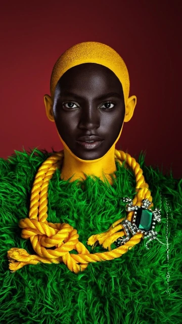 Portrait of an African American woman with yellow paint on her face and a rope necklace with a jeweled pendant, set against a red backdrop.