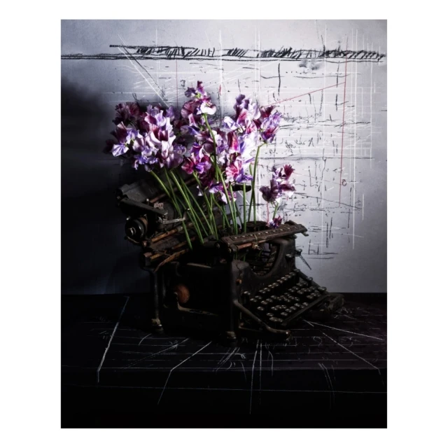 Antique black typewriter overflowing with purple and white flowers, set against a textured gray and white artistic background.