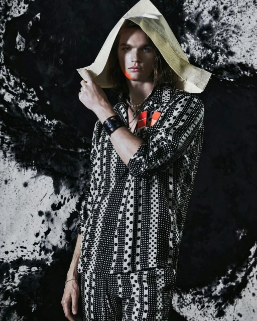 Young man in a patterned shirt and pants poses against a dark, splattered background, wearing a makeshift hat and accessories.