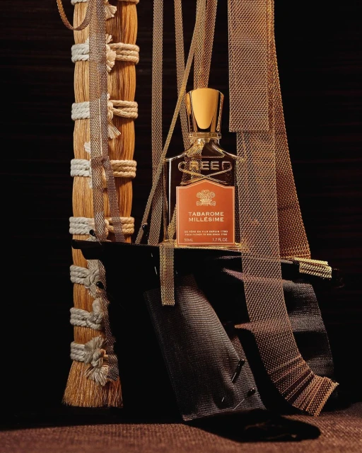 A Creed Tabarome Millesime perfume bottle is the central focus, adorned with gold ribbons and positioned on a dark surface.