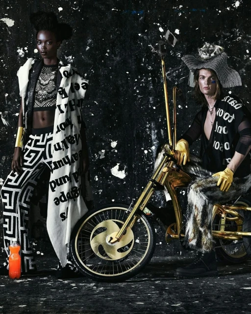 Two fashion models wearing bold patterned clothing posing next to a gold motorcycle against a black and white mottled backdrop in a studio.
