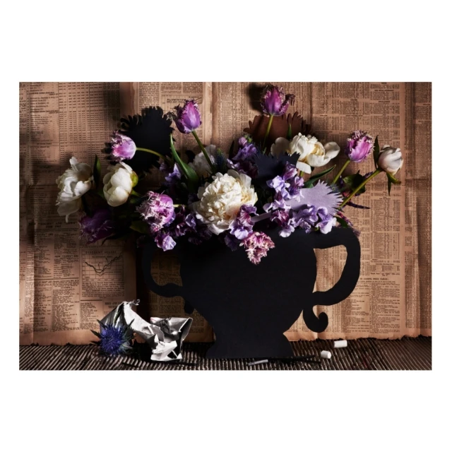 Still life photo of a black vase filled with purple and white flowers, set against a backdrop of vintage newspaper and wood.