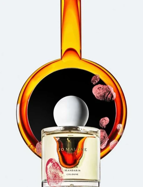 Jo Malone cologne bottle close-up with abstract amber perfume background, white cap, and orange accents on a white background.